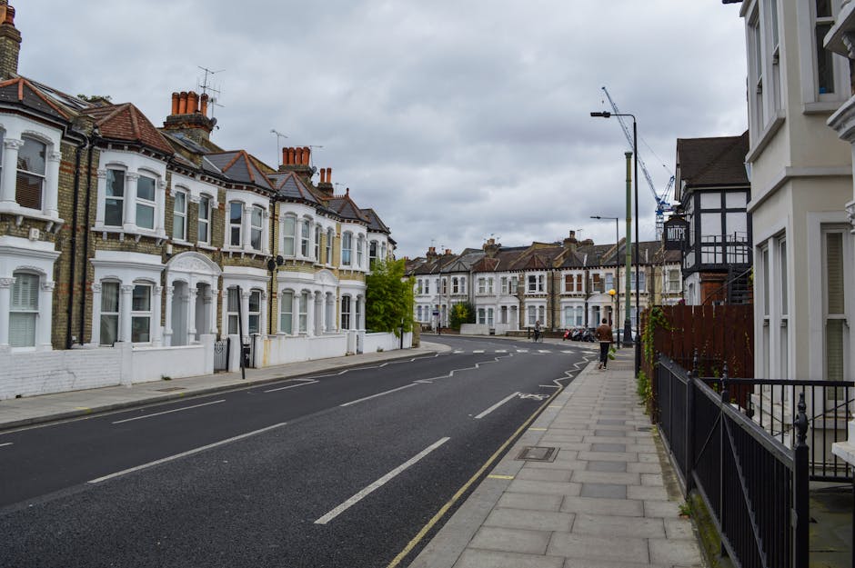 A suburban street scene viewed from the sidewalk on the right, with a row of Victorian-style terraced houses featuring white facades, bay windows, decorative mouldings, and brick chimneys. The houses are set back slightly from the road, with small front gardens or steps, and some have black metal fences. The street is paved with asphalt, with white dashed lines and parking bays along the curb. A construction crane is visible in the distance on the right side, suggesting ongoing building work. A man is walking away from the camera on the pavement, carrying a box, possibly part of a home removal or moving process. To the right, behind a black metal fence, a white house with a bay window and a side door is partially visible. Overcast skies with grey clouds cast diffuse lighting over the scene. This image illustrates a typical residential area suitable for house removals, with a focus on the street environment and nearby properties, reflecting the services offered by Man With a Van Kennington for house relocations and furniture transport.