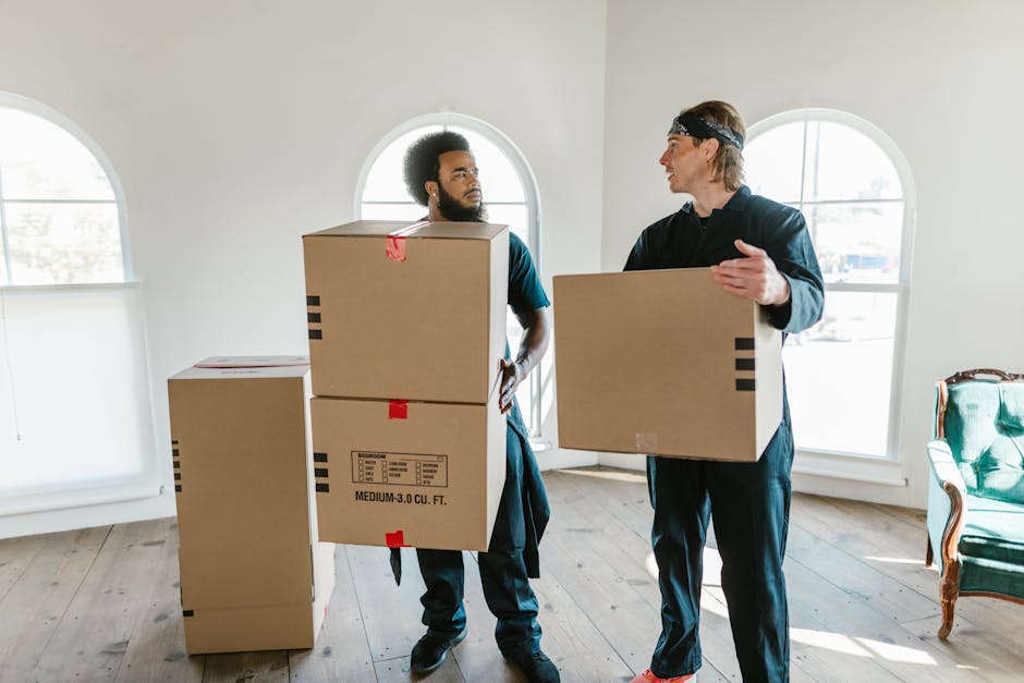 Two men are inside a well-lit room with large arched windows that allow natural light to illuminate the space. They are engaged in a home relocation process, with one man holding a medium-sized cardboard box labeled with handling instructions and capacity details, while the other man carries a similar box. The room has a light-colored wooden floor and minimal decor, with a green upholstered armchair positioned near the window on the right. Behind the men, there are several other cardboard boxes, some stacked, prepared for loading or unpacking. The men are dressed casually; one is wearing dark trousers and a matching jacket, and the other has a headband and a dark top, both appearing focused on their task. The scene captures a moment of packing and furniture transport as part of a professional removal service, where careful handling and organized loading are evident.