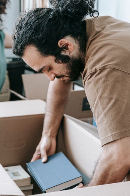 A man with curly hair and a beard, dressed in a blue shirt and dark trousers, sitting on the wooden floor of a room next to several cardboard moving boxes sealed with red packing tape. The boxes are s