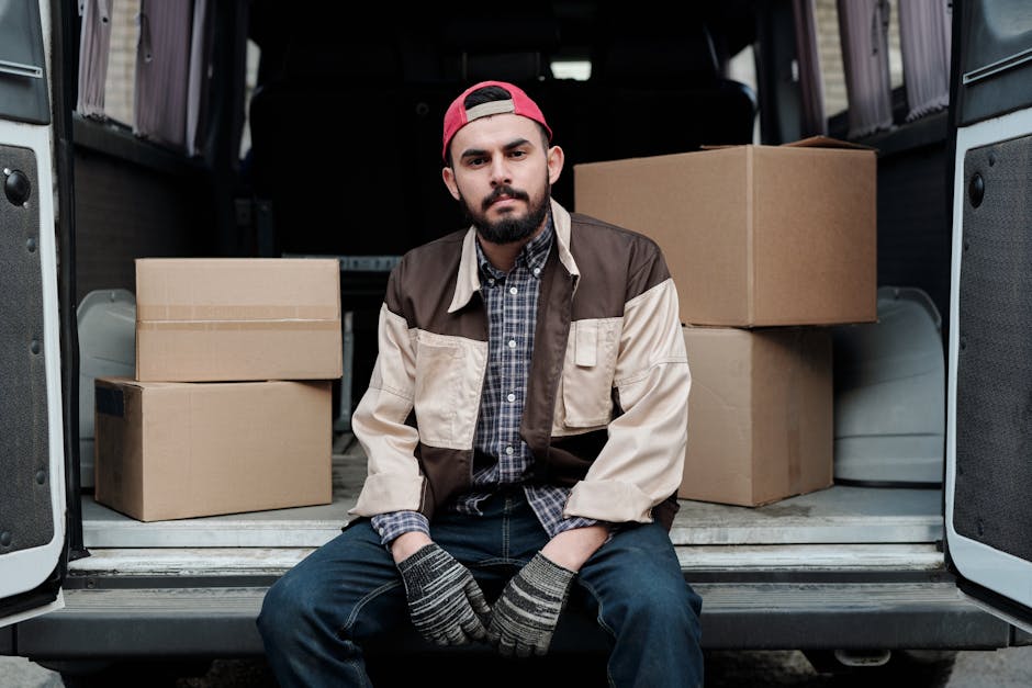 A man stands outside a residential property next to a large white van with its rear doors open, preparing for a home relocation. He is holding a cardboard box and is about to place it among several ot