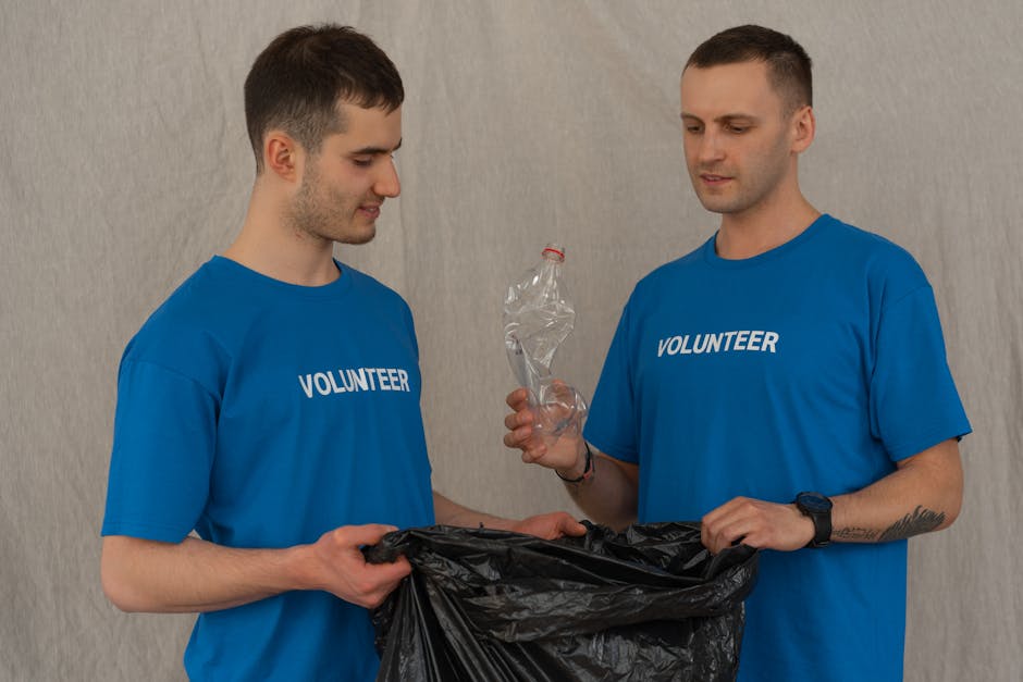 Two male volunteers wearing blue T-shirts with the word 'VOLUNTEER' printed in white are participating in a home relocation or community cleanup activity, holding a black plastic trash bag with one ma