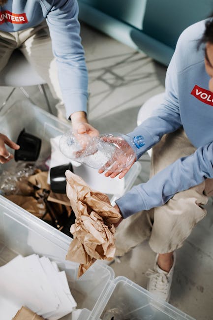 Two male volunteers wearing blue T-shirts with the word 'VOLUNTEER' printed in white are participating in a home relocation or community cleanup activity, holding a black plastic trash bag with one ma