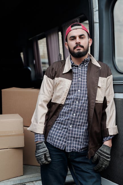 A man wearing a black cap, yellow vest, and grey trousers is in the process of loading a medium-sized cardboard box into the open rear of a white panel van parked on the street outside a residential p